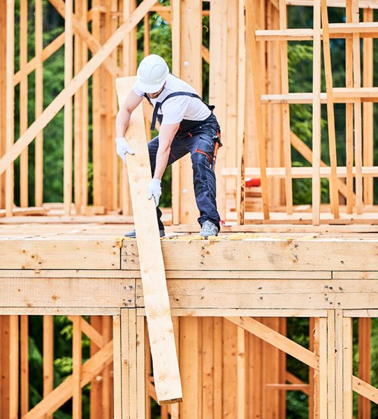 Carpenter holding large beam while builds wooden frame house near the forest.